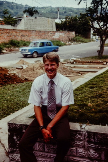 John Quist as a young man, seated outdoors in a white shirt and tie, smiling